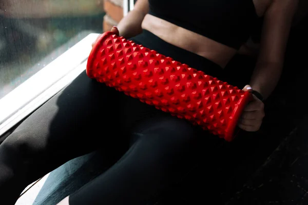 Primer plano de la mujer joven en un uniforme deportivo negro se encuentra después del entrenamiento con un cilindro de masaje rojo para trabajar la fascia de los músculos del cuerpo —  Fotos de Stock