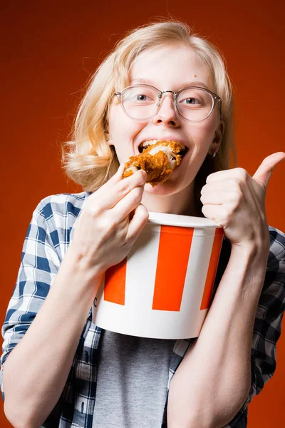 Atraktivní blondýnka s brýlemi drží vědro rychlého občerstvení a jí kuřecí stehno, raduje se na konci stravy na červeném pozadí — Stock fotografie
