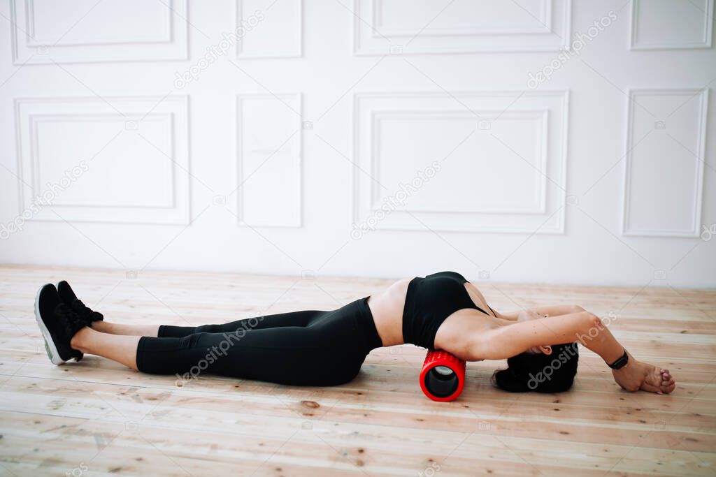 Asian young woman doing back and hip muscles strengthening exercise with fascia massage roller lying on floor in classic interior