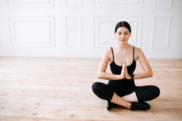 Young meditating woman in black sportswear holding hands in front of her and sitting on wooden floor in lotus position