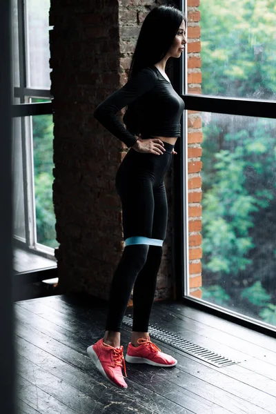Κορίτσι που εκπαιδεύει τους μυς των ποδιών χρησιμοποιώντας ένα ελαστικό λάστιχο expander στέκεται μπροστά από ένα πανοραμικό παράθυρο σε μια σοφίτα — Φωτογραφία Αρχείου