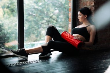 Atletik Asyalı kız yerde oturuyor ve elinde pilates tüpüyle büyük bir pencereden dışarı bakıyor.