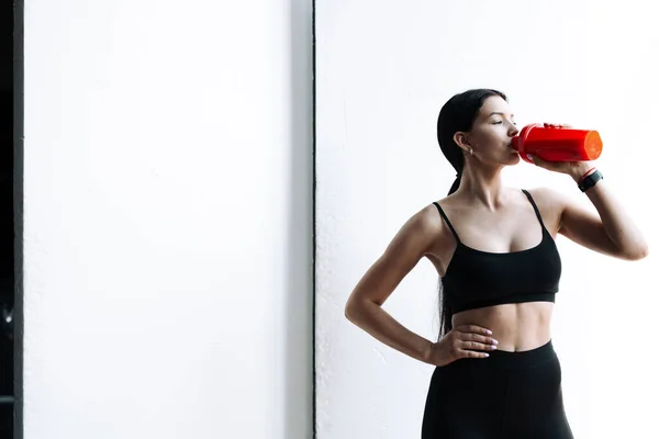 Foto horizontal de una deportista asiática en un top negro y polainas que bebe agua de una botella roja sobre un fondo blanco —  Fotos de Stock