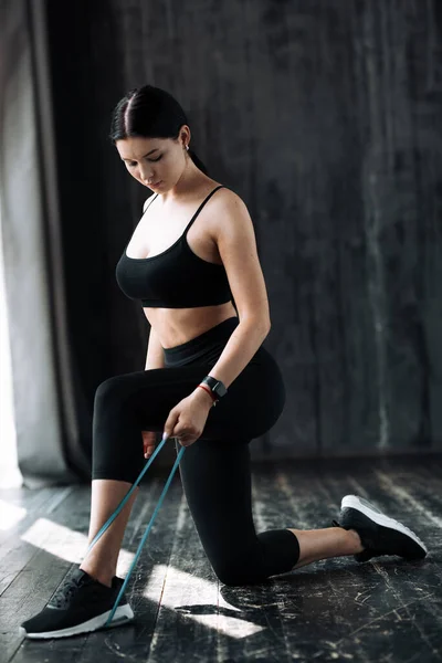 Flickan är på knä och trycker en elastisk expander med benet för att bygga upp musklerna i armarna — Stockfoto