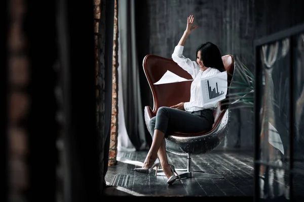 Girl office worker sits in a chair in the office, rejoices and throws documents into the air