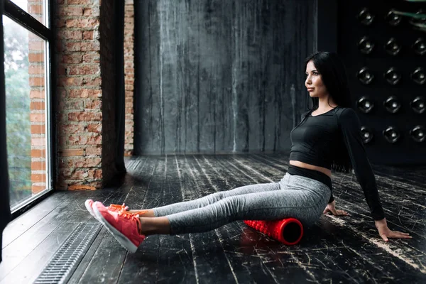 Una chica morena en un chándal gris se sienta en un cilindro de masaje rojo para resolver la fascia y mira por la ventana — Foto de Stock