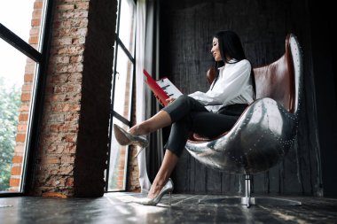 Aspen sandalyesinde oturan ve elinde belgelerle kırmızı bir dosya tutan bir kızın profili.