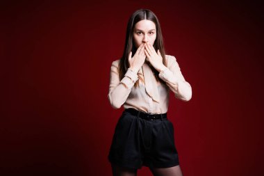 Esmer kız kırmızı arka planda duruyor ve elleriyle ağzını kapatıyor.