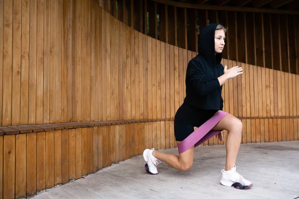 Menina em preto esportes uniforme lunges com uma faixa de borracha fitness — Fotografia de Stock