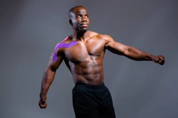 Hombre musculoso de piel oscura con un yeso para kinesioterapia se levanta sobre un fondo gris — Foto de Stock