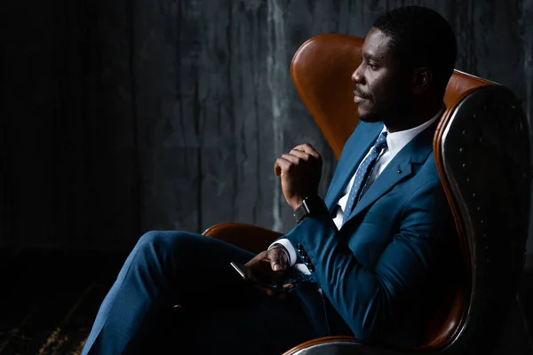 Hombre de negocios afroamericano en un traje clásico azul se sienta en un sillón de aviador loft con un teléfono en sus manos —  Fotos de Stock