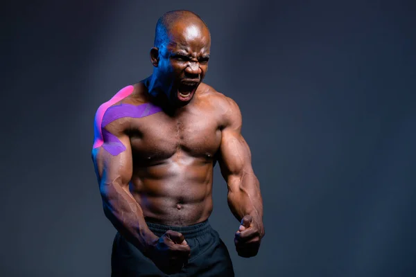 Un hombre afroamericano se para sobre un fondo gris y tensa sus músculos con un yeso de kinesioterapia en la articulación de su hombro — Foto de Stock