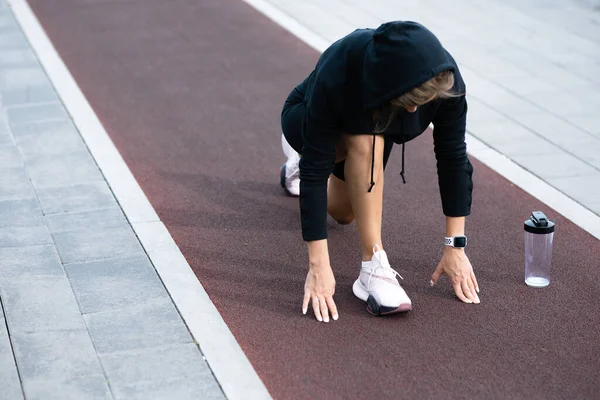 Una chica con un uniforme deportivo negro se encuentra en una posición de partida baja en una cinta de correr — Foto de Stock