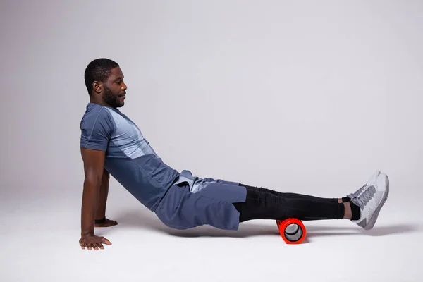 Hombre afroamericano haciendo ejercicio con rollo de masaje sobre fondo blanco. Obras en la fascia de la parte posterior del muslo —  Fotos de Stock