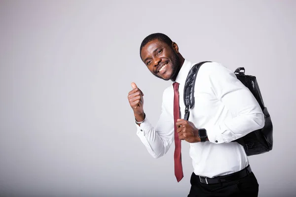 Αφρο-Αμερικανός φοιτητής με λευκό πουκάμισο και με μαύρο σακίδιο στον ώμο στέκεται σε λευκό φόντο, χαμογελάει και σηκώνει τον αντίχειρά του. — Φωτογραφία Αρχείου