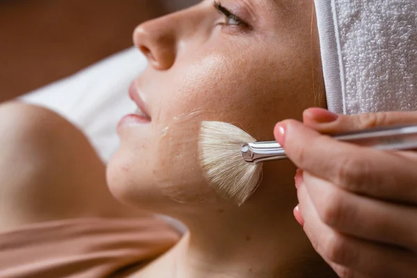 Primer plano de la cara de una mujer con una toalla blanca en la cabeza. Manos con un cepillo para aplicar la máscara. Cuidado de la piel y cosmetología — Foto de Stock