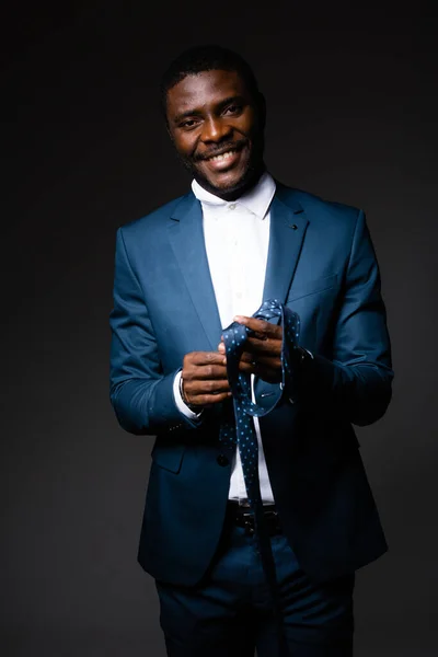 Ένας άνδρας επιχειρηματίας με ένα μπλε κλασικό κοστούμι στέκεται σε ένα γκρι φόντο και κρατά μια γραβάτα στα χέρια του — Φωτογραφία Αρχείου