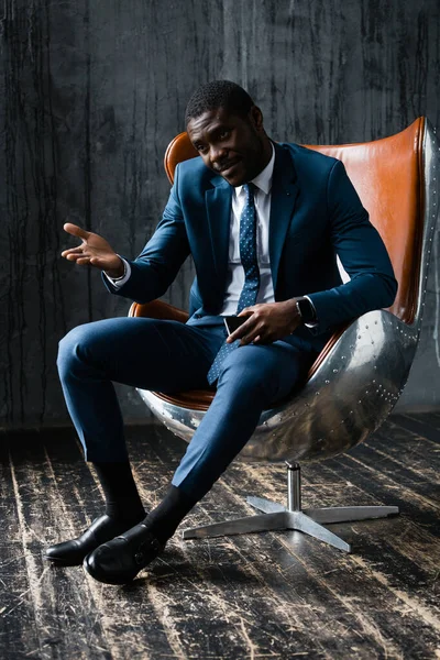 Um homem afro-americano num fato clássico azul senta-se numa cadeira de aviador, olha para a câmara e aponta com a mão para algo — Fotografia de Stock