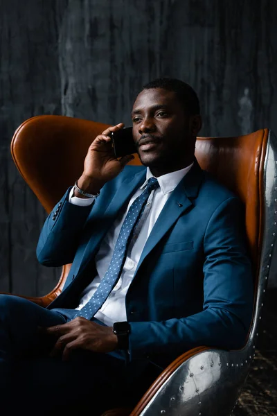 Retrato de um homem de pele escura sentado em um terno azul clássico em uma cadeira de escritório e falando ao telefone — Fotografia de Stock