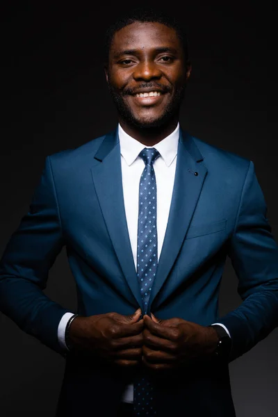 Ένας Αφροαμερικάνος στέκεται με ένα μπλε κλασικό κοστούμι, προσαρμόζει το γιακά με τα χέρια του και χαμογελάει. — Φωτογραφία Αρχείου