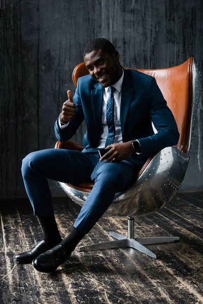 Homem de negócios negro em um terno clássico senta-se em uma cadeira de escritório, sorri e levanta o polegar — Fotografia de Stock