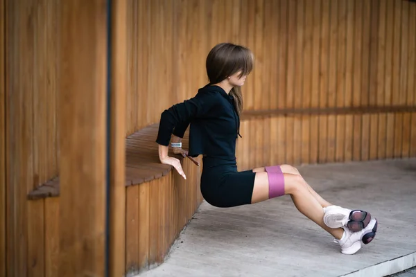 Una chica con un uniforme deportivo negro hace flexiones inversas con una banda elástica para entrenar para entrenar tríceps — Foto de Stock