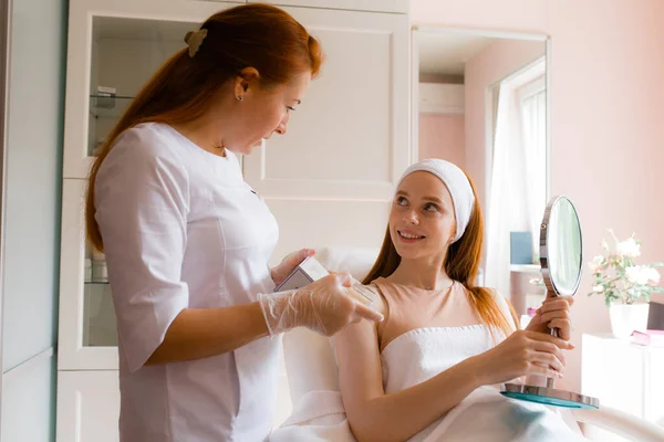 Doctor beautician talking to a red-haired girl who sits on a white medical couch and holds a mirror in her hands — Stock Photo, Image