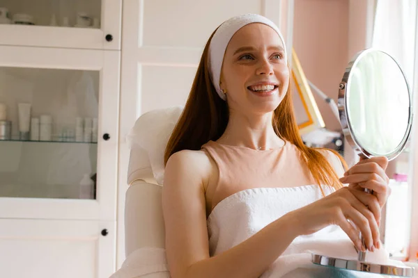 한 소녀가 머리에 흰 붕대를 감고 미용사의 사무실에 앉아 손에 거울을 들고 미소를 짓습니다 — 스톡 사진