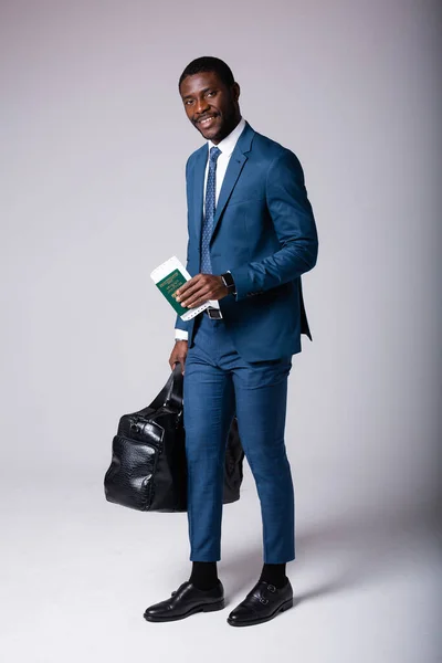Αφροαμερικάνος με μεγάλη μαύρη ταξιδιωτική τσάντα και διαβατήριο στα χέρια στέκεται σε γκρι φόντο — Φωτογραφία Αρχείου
