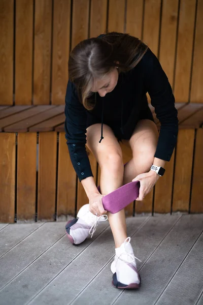 La chica se está preparando para el entrenamiento con un expansor elástico. Se sienta en un banco y le pone un elástico de entrenamiento en la pierna. — Foto de Stock