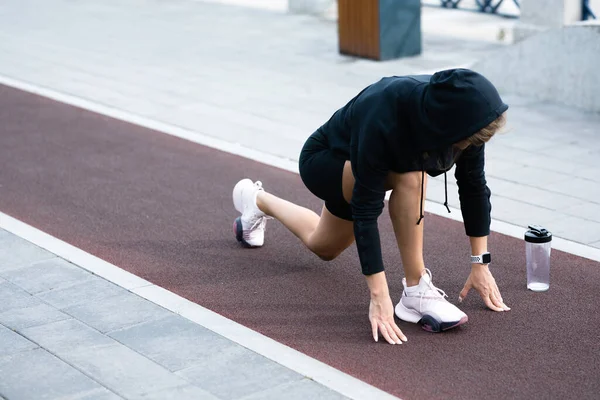 Una chica con un uniforme deportivo negro se encuentra en una posición de partida baja en una cinta de correr — Foto de Stock