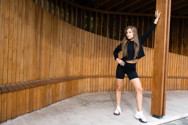 Chica en ropa deportiva negra para dar un paseo por el parque. Se apoya en un poste de madera en un parque de recreo —  Fotos de Stock