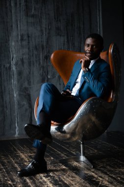 Mavi takım elbiseli bir ofis çalışanı tavan arası bir uçuş koltuğunda oturur, elinde bir telefon tutar ve ciddi bir yüzle kameraya bakar.