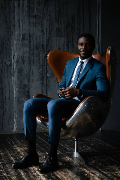 Ένας άνδρας επιχειρηματίας με ένα μπλε κλασικό κοστούμι κάθεται σε μια καρέκλα πατάρι αεροπόρος σε ένα γκρι φόντο τοίχο — Φωτογραφία Αρχείου