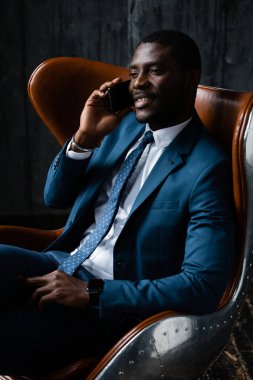 Koyu tenli bir adamın portresi klasik mavi bir takım elbiseyle bir ofis koltuğunda oturmuş telefonda konuşuyor.