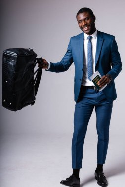 Siyah seyahat çantası ve pasaport taşıyan, gülümseyen ve gri arka planda duran mavi takım elbiseli Afrikalı bir adam.