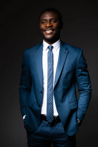 Bonito homem americano africano em azul terno clássico fica no fundo escuro e sorri — Fotografia de Stock