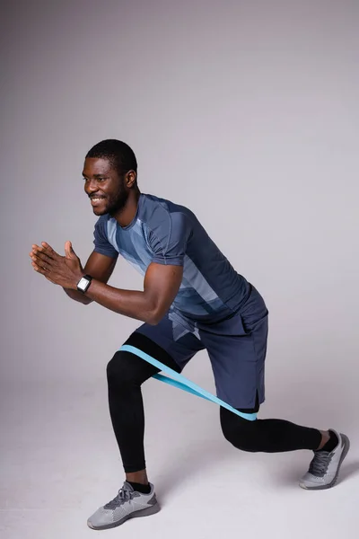 Entrenador de fitness afroamericano se abalanza con elástico de entrenamiento azul. Muestra ejercicio sobre un fondo gris —  Fotos de Stock