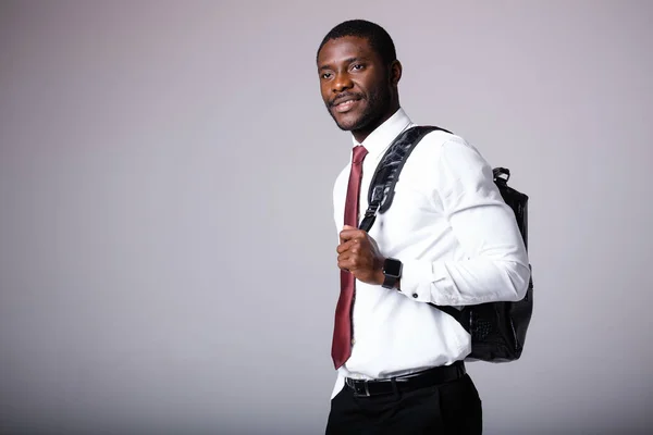 Um homem afro-americano de camisa branca com uma mochila preta no ombro está em um fundo cinza. Programador jovem empregado — Fotografia de Stock