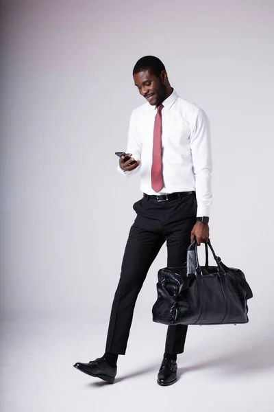 Um homem de negócios afro-americano está em um saco de viagem grande e segura um telefone em suas mãos. Conceito de compra online de bilhetes de avião — Fotografia de Stock