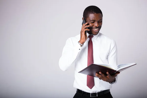 Šťastný Afroameričan v bílé košili stojí na šedém pozadí s otevřenou složkou a mluví po telefonu. Uzavře smlouvu s klientem telefonicky — Stock fotografie