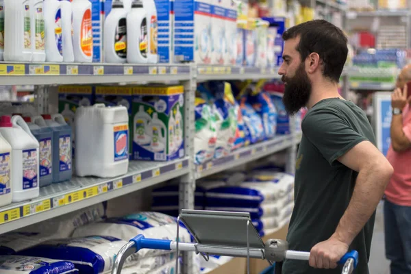 Süpermarkette sakallı genç adam el arabasını tutarken bir rafta deterjan görüyor.