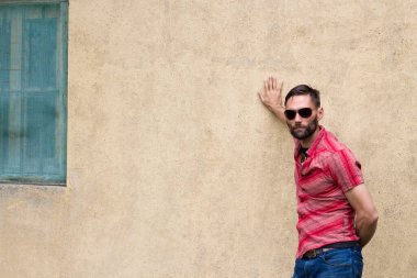 Güneş gözlüklü ve sakallı bir adam eski bir evin duvarına yaslanmış ve kameraya bakıyor.