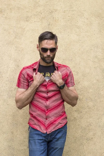 Man with a beard and sunglasses posing with shirt and jeans with chain, like a rock star. He is with serious attitude and grabs the shirt collar with both hands