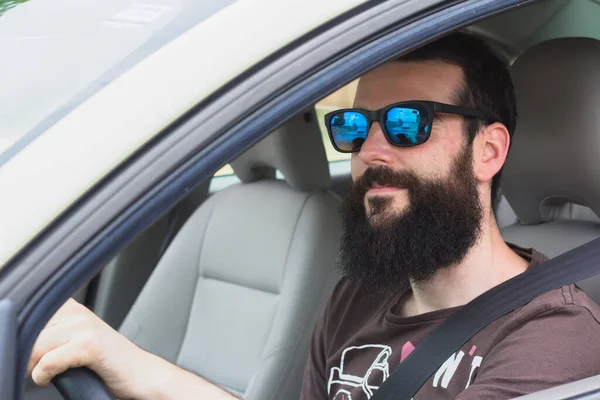 Sakallı ve güneş gözlüklü genç adam direksiyonu tutuyor.