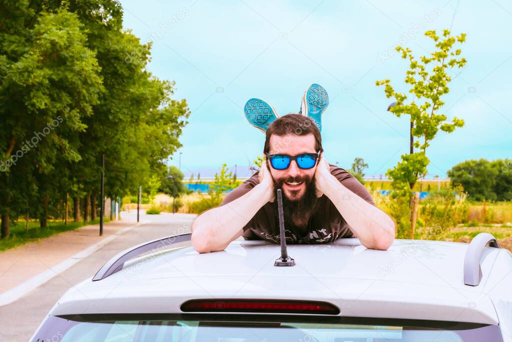 Holidays concept. Young man with beard and sunglasses lying on the roof of a car. He is smiling and looking to the camera.