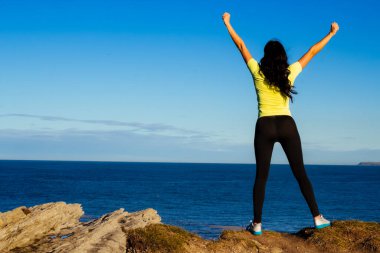 Başarılı sporcu kadının, okyanusun önündeki mavi gökyüzüne kollarını kaldırışının arka planı. Kolları havada spordan sonra gol atmayı kutlayan sporcu. Boşluğu kopyala