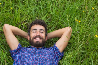 Mutlu genç adam gülümsüyor ve çimenlerde uzanıyor. Elleri başının arkasında kameraya bakıyor.