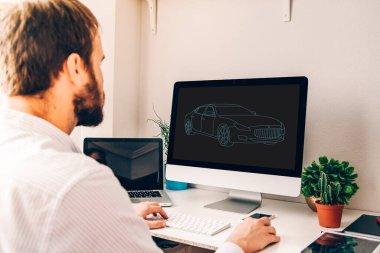 Bilgisayarla çalışan genç bir adam. Ekranda araba çizimi var. Tablet, laptop, akıllı telefon ve bitkilerle dolu bir masada.