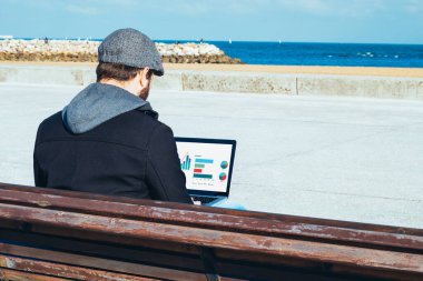 Bilgisayarında iş grafikleri olan genç bir adam plajın önündeki bankta oturuyor.